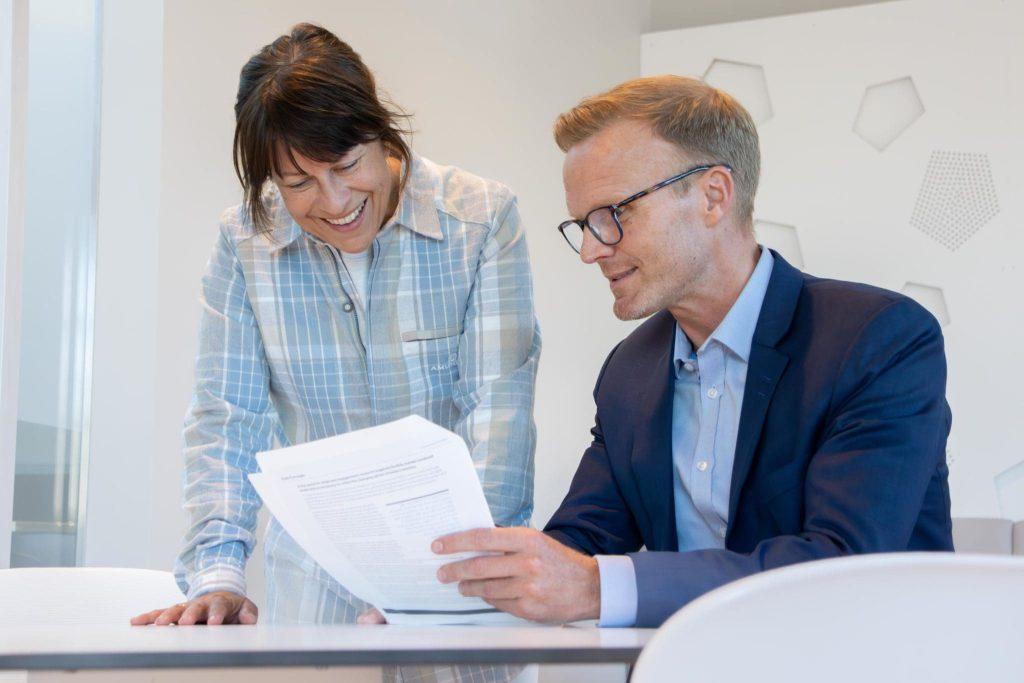 En dame står og lener seg på et bord. En mann i dress sitter ved bordet og holder opp et ark. De ser begge på arket og smiler.